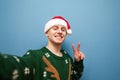Smiling young man in santa hat and green sweater makes christmas selfie on blue background, shows to the camera a gesture of peace Royalty Free Stock Photo