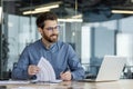 A smiling young man sits in the office at a table in front of a laptop, makes notes in documents, works with data Royalty Free Stock Photo