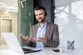 Smiling young man sitting at desk in office wearing headset and talking gesturing hands on video call via laptop Royalty Free Stock Photo