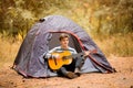 Smiling young man sitting near touristic tent and playing guitar in forest Royalty Free Stock Photo