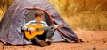 Smiling young man sitting near touristic tent and playing guitar in forest Royalty Free Stock Photo