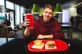 Smiling young man is sitting at the table with a fast food tray holding a glass of drink in his hand Royalty Free Stock Photo