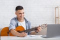 Smiling young man watching online lesson, playing guitar and makes notes