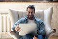 Smiling young man using laptop, sitting in cozy armchair