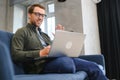 Smiling young man wearing glasses using laptop, sitting in cozy armchair, happy male looking at computer screen Royalty Free Stock Photo