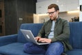 Smiling young man wearing glasses using laptop, sitting in cozy armchair, happy male looking at computer screen Royalty Free Stock Photo