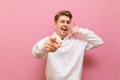 Smiling young man in white sweatshirt shows call me gesture isolated on pink background. Positive guy points his finger at the Royalty Free Stock Photo