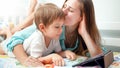 Smiling young mother kissing her little son lying in bed and watching video on digital tablet computer Royalty Free Stock Photo