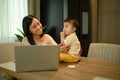 Smiling young mother working remotely on laptop while taking care of baby son on dinning table Royalty Free Stock Photo