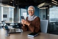 Smiling young Muslim woman in hijab working in office on laptop, using mobile phone, smiling at camera Royalty Free Stock Photo