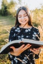 Smiling young religious girl reading her bible, outside in the field at sunset. Spiritual revival.