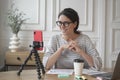 Smiling young spanish businesswoman in glasses sitting in front of smartphone on tripod at workplace