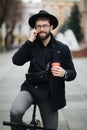 Smiling young stylish bearded hipster man using mobile phone while drinking coffee outdoors with bicycle Royalty Free Stock Photo
