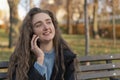 Smiling young woman in autumn park with wavy brown hair sits on bench in park talks on phone and gesticulating Royalty Free Stock Photo