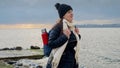 Smiling young woman with a backpack and thermos walking along a rocky sea beach, braving the cold and windy weather in pursuit of