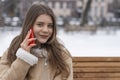 Smiling young woman in beige sheepskin coat rests outside in winter. Cute girl sits on park bench and talks on phone Royalty Free Stock Photo