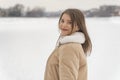 Smiling young woman in beige sheepskin coat standing half turn on snowy forest background. Winter outdoor walking Royalty Free Stock Photo