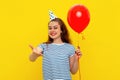 Smiling young woman in a birthday cap holds red inflated helium balloon and reach hand to the camera, celebrates birthday or Royalty Free Stock Photo