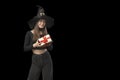 Smiling young woman in black pointed hat holds a gift. Looks at camera, gift box. Witch girl isolated on black background. Copy Royalty Free Stock Photo