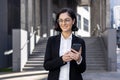 Smiling young woman in a business suit outside an office center, holding a phone in her hands and looking sullenly to Royalty Free Stock Photo