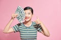 Smiling young woman closes half of her face with a fan of banknotes cash and shows thumb up on pink background