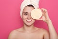 Smiling young woman with cosmetic sponge in hands, standing and covering one eye isoalted over pink studi obackground, beautiful