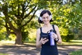 Smiling young woman doing sports and jogging outdoors in park, standing resting in headphones, holding water bottle and Royalty Free Stock Photo