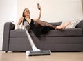 smiling young woman in elegant long black dress is sitting on sofa and using cellphone holding vacuum cleaner Royalty Free Stock Photo