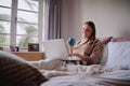 Smiling young woman enjoying morning coffee using laptop sitting in her bed working from home - Student studying online