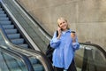Smiling young woman on escalator, talking on mobile phone, drinking coffee and looking happy, going to university or Royalty Free Stock Photo