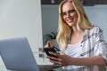 smiling young woman in eyeglasses holding smartphone and using laptop while working