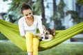 Smiling young woman in green hammock with cute dog Welsh Corgi in a park outdoors. Beautiful happy female in white shirt Royalty Free Stock Photo