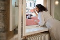 Smiling young woman greeting someone with her hand while looking out the window and enjoying her morning coffee Royalty Free Stock Photo