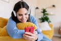 Smiling young woman with headphones using mobile phone lying on sofa at home Royalty Free Stock Photo