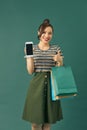 Smiling young woman hold package bag with purchases pointing index finger on mobile phone isolated on green background. Shopping