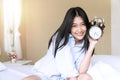 Smiling woman holding alarm clock sitting on the white bed while looking at camera. Cheerful girl waking up in the morning Royalty Free Stock Photo