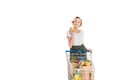 smiling young woman holding apple and looking away while standing with shopping trolley with grocery bags