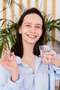 Smiling young woman holding pill omega capsule and glass of water indoors. Girl taking medicine at home Royalty Free Stock Photo