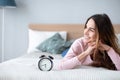 Smiling young woman lying awake in bed with alarm clock, good morning