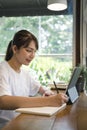 Smiling young woman making note on notebook and using computer tablet at coffee shop. Royalty Free Stock Photo