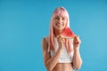 Smiling young woman with natural long pink dyed hair holding a slice of watermelon, posing isolated over blue studio Royalty Free Stock Photo