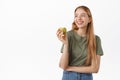 Smiling young woman with perfect white teet, looking aside and holding apple, eating fruit and thinking, standing over Royalty Free Stock Photo
