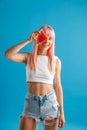 Smiling young woman with pink hair holding a slice of watermelon over her eye, standing  over blue studio Royalty Free Stock Photo