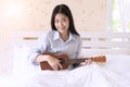 Smiling young woman playing Ukulele relax on the white bed in the morning. Joyful girl play music with ukulele in bedroom Royalty Free Stock Photo