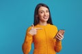 Smiling young woman pointing at smartphone, wearing orange sweater over blue Royalty Free Stock Photo