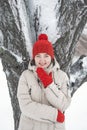 Smiling young woman in red hat and mittens stands near snow-covered tree. Portrait of cute thoughtfully girl looking to the side