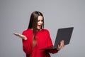 Smiling young woman in red sweater holding laptop computer and pointing at copyspace while looking at the camera over gray Royalty Free Stock Photo