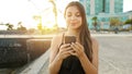 Smiling young woman sending message with smart phone at sunset with seaside town on the background. Positive trendy girl blogging Royalty Free Stock Photo