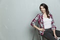 Smiling young woman sits on a stepladder against the background of a gray wall. Beautiful brunette in jeans and a plaid shirt. Royalty Free Stock Photo
