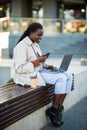 Smiling young african woman sitting on bench outdoors at the street, using laptop , talking on mobile phone Royalty Free Stock Photo